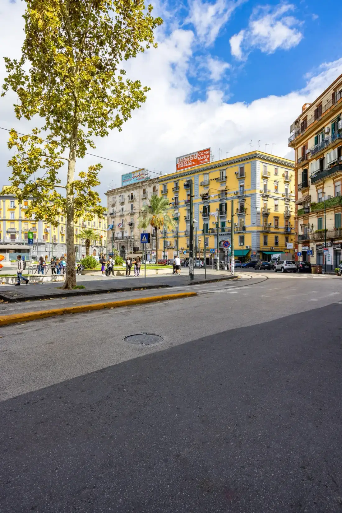 Una calle y cruce urbano con peatones, árboles y edificios de varios pisos.