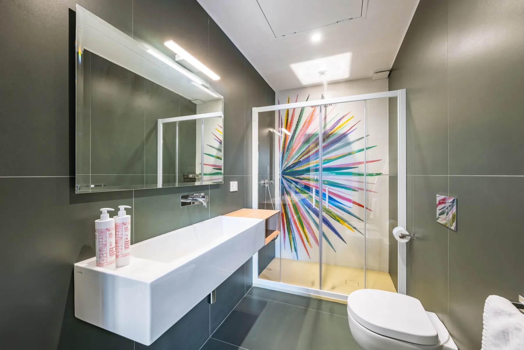 Un baño moderno con un lavabo flotante y una obra colorida en la mampara de la ducha.