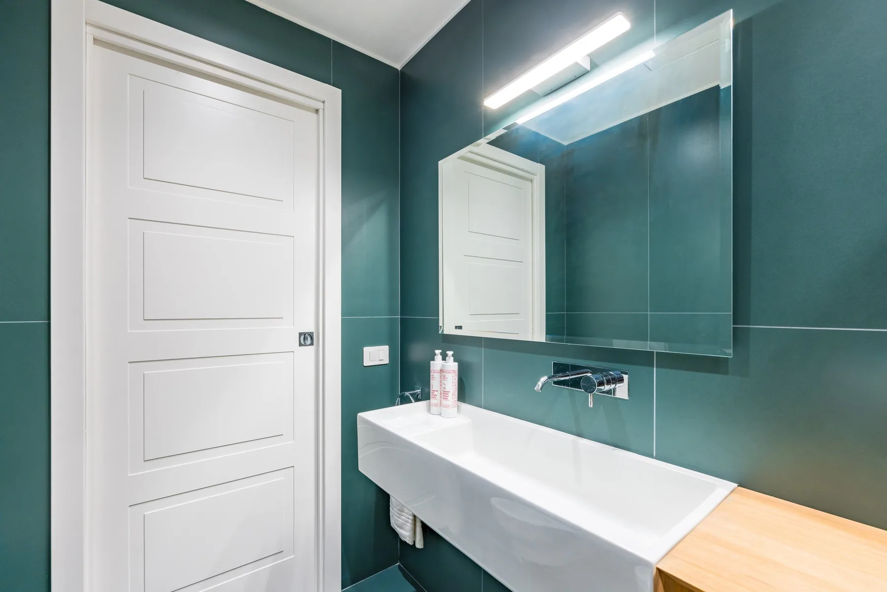 Un baño con lavabo blanco, espejo grande y puerta blanca.
