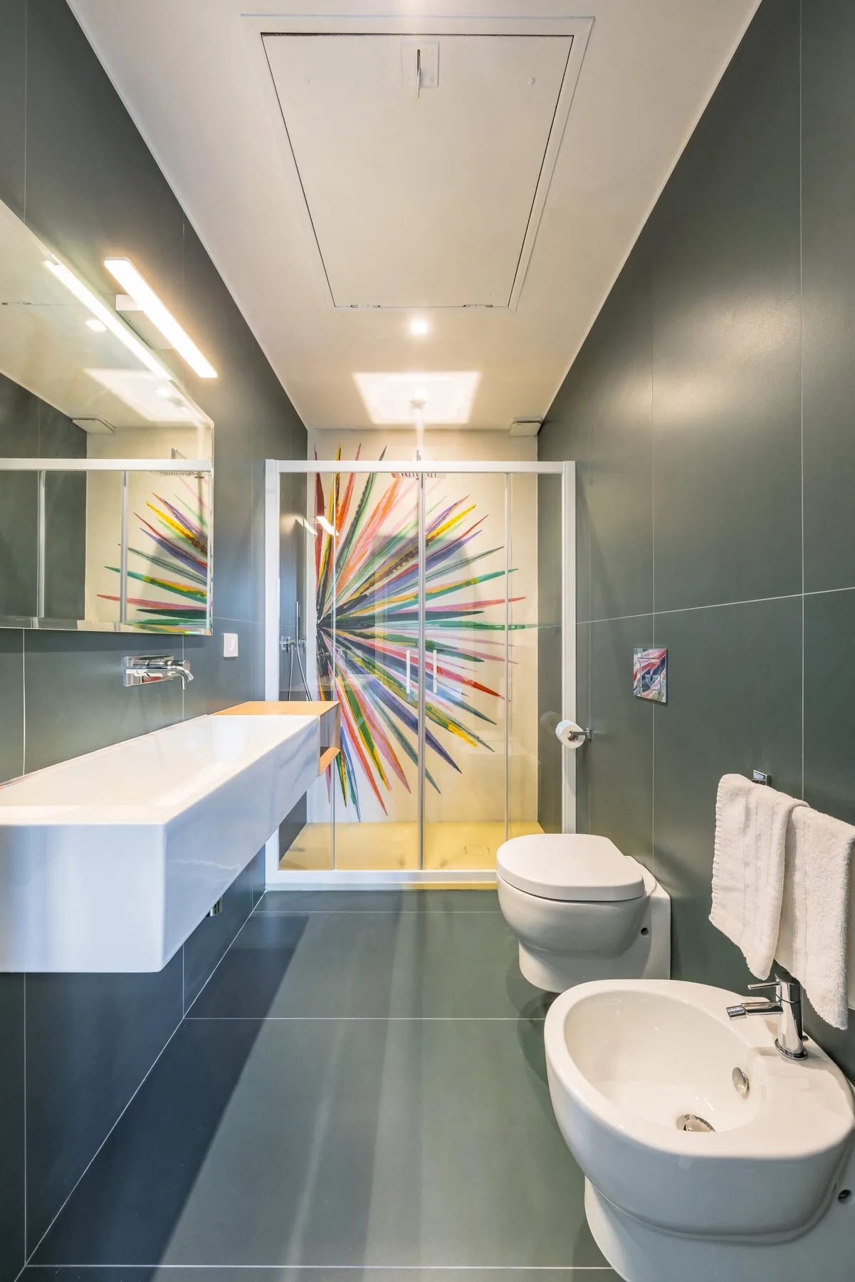 Un baño moderno con lavabo suspendido, inodoro y una obra de vidrio colorida.