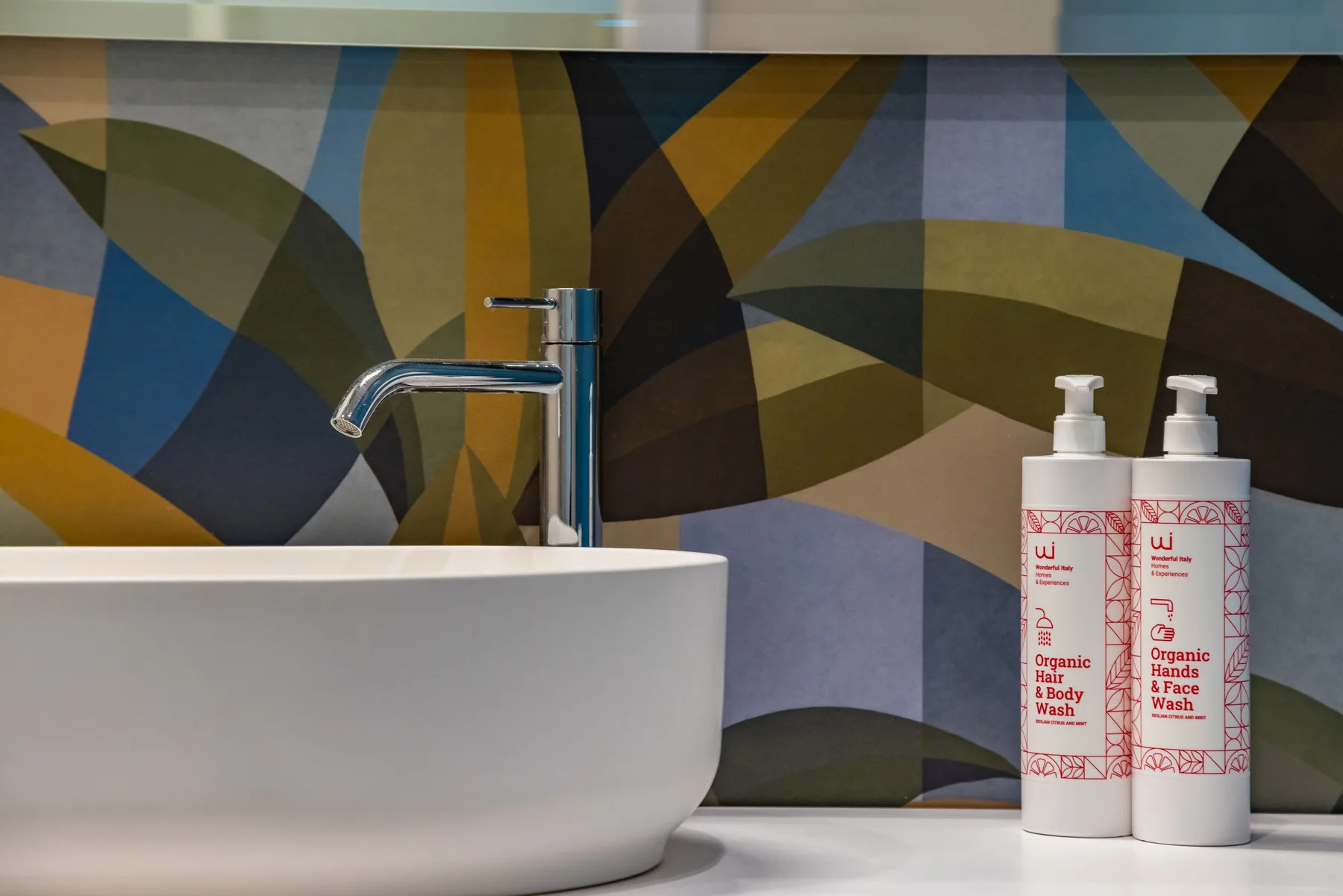 Un lavabo con grifo está frente a una pared geométrica colorida, junto a dos botellas con dispensador.