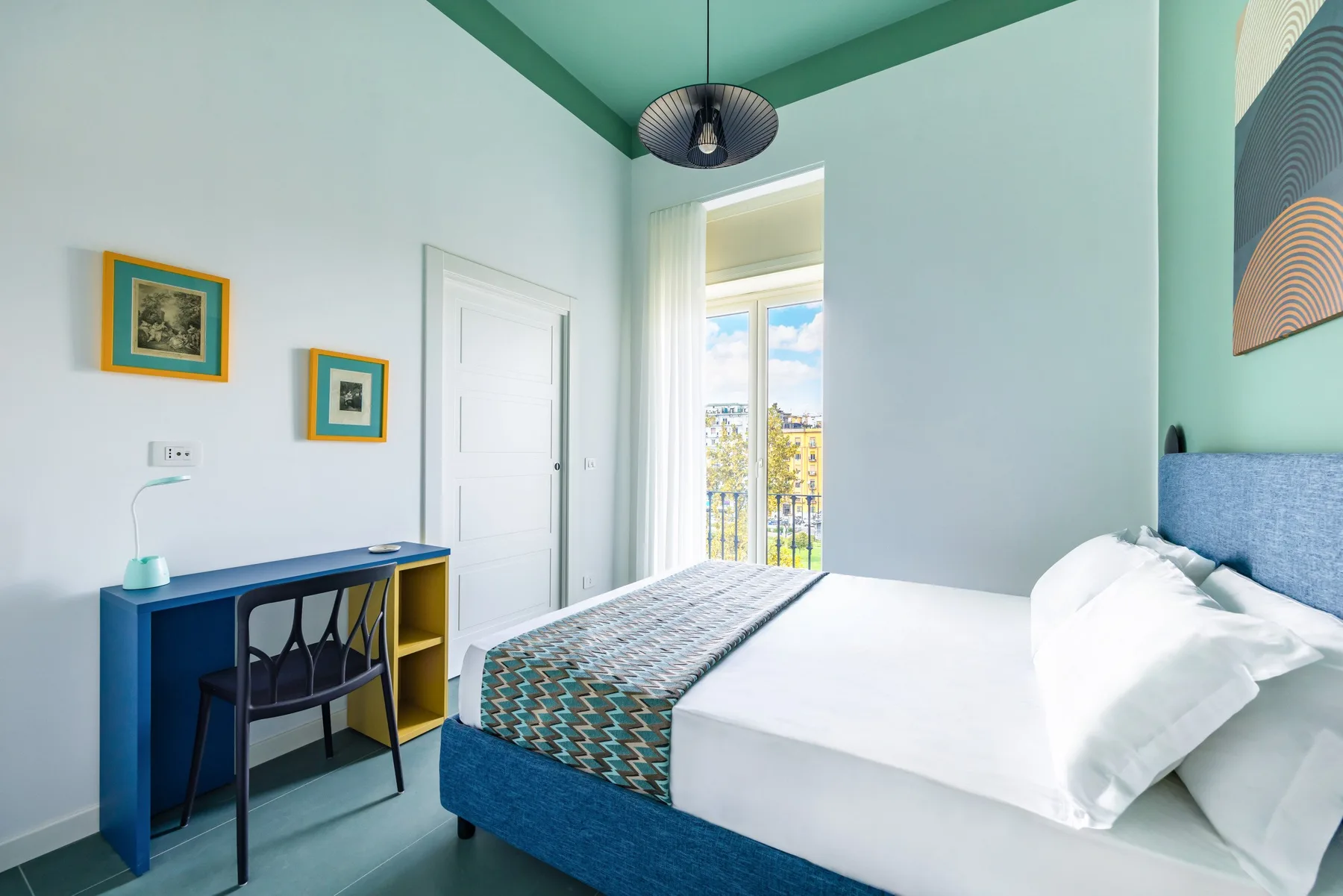Un dormitorio con una cama, un escritorio y una ventana alta que da a un balcón.