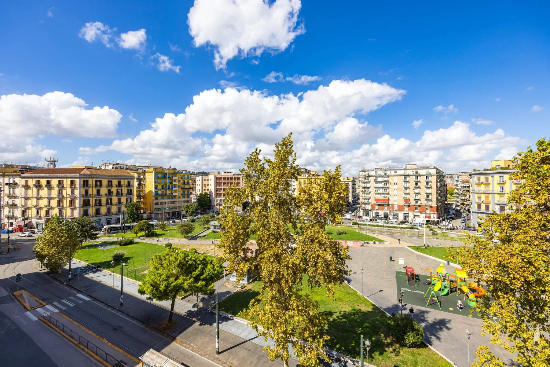 Un parque urbano soleado con edificios de apartamentos y una zona de juegos infantil.