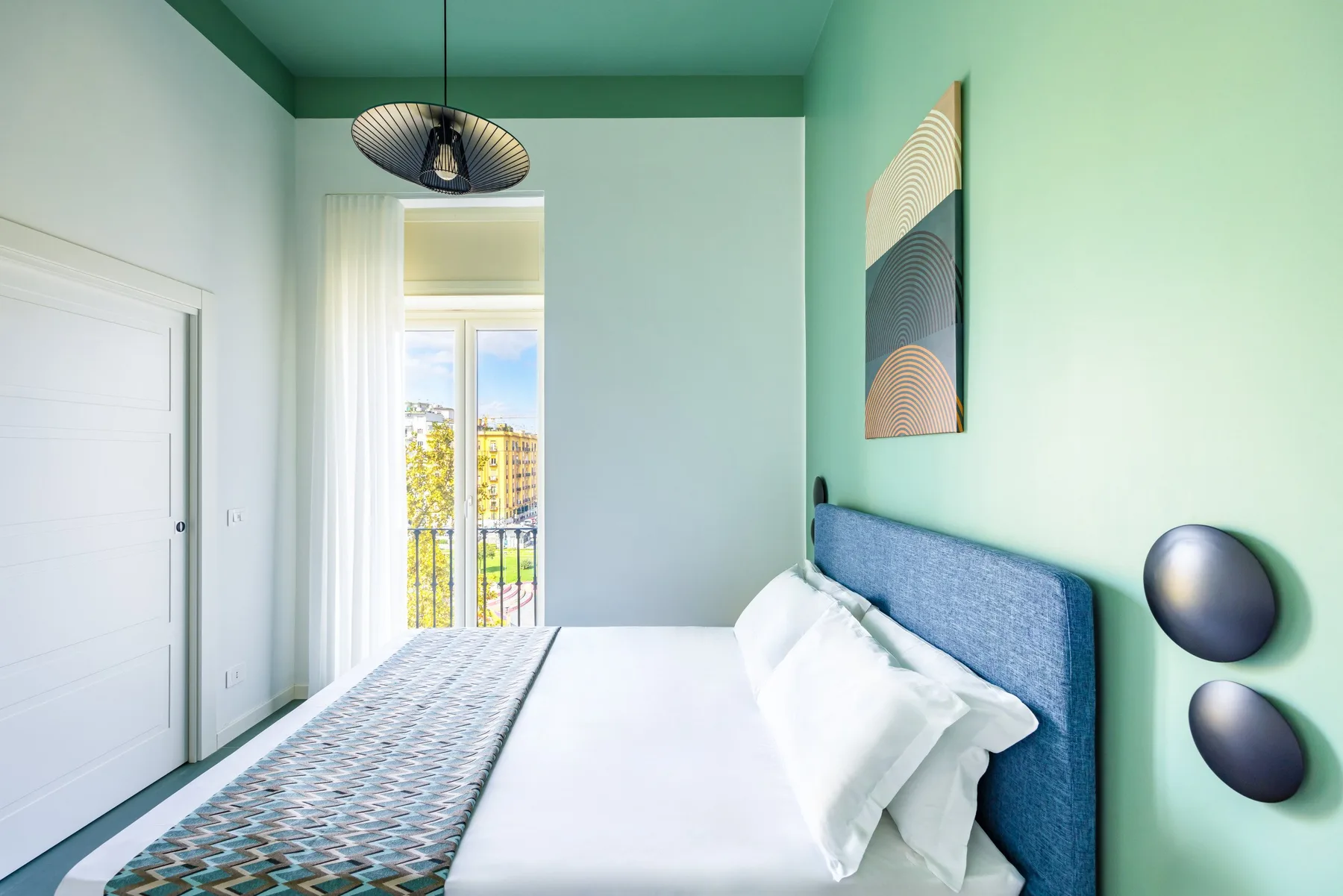 Un dormitorio luminoso con una cama junto a una ventana alta y ropa de cama estampada.