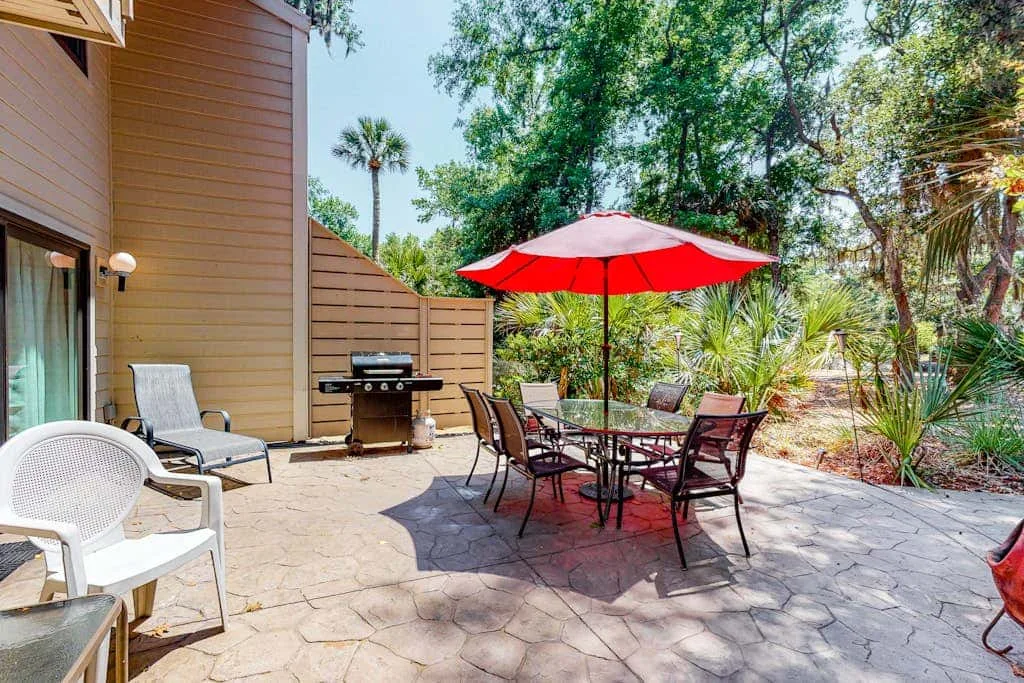 An outdoor patio has a dining table beneath a red umbrella and seating nearby.