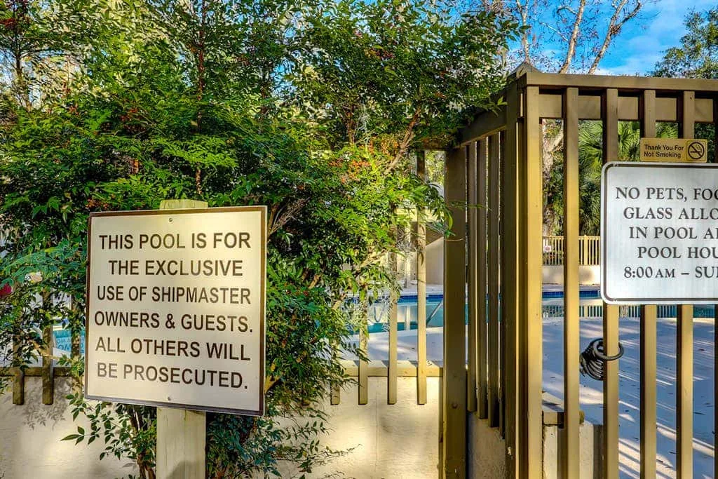 A gated outdoor swimming pool is surrounded by trees and posted notices.