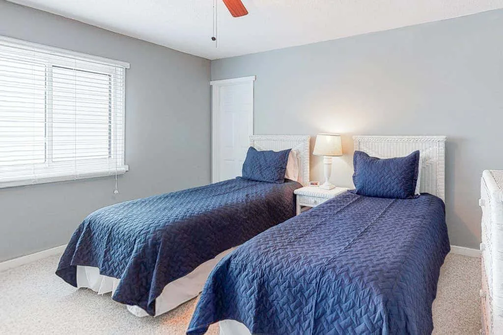 A bedroom with two twin beds covered in blue bedspreads beside a window.