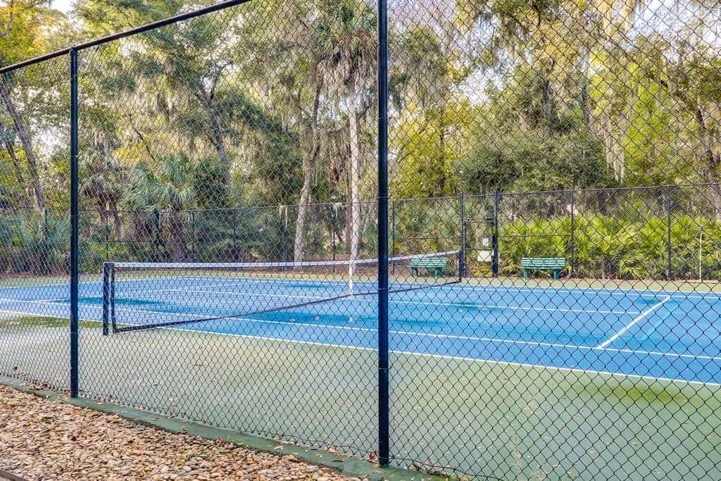 An outdoor tennis court is enclosed by a chain-link fence, with trees behind it.
