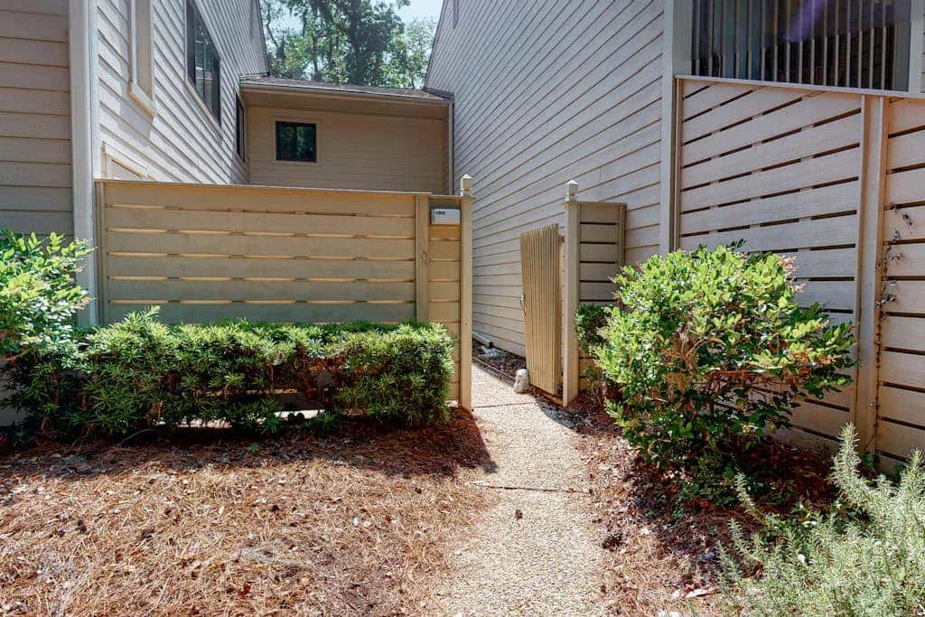 A narrow outdoor walkway runs between buildings beside wooden privacy screens and shrubs.