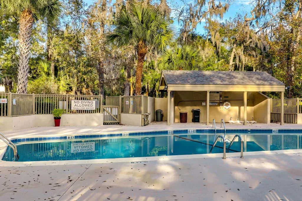 An outdoor swimming pool sits beside a covered poolside area amid tall trees.