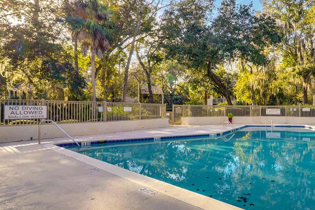 An outdoor swimming pool sits beside a fenced deck beneath leafy trees.