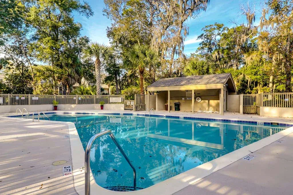An outdoor swimming pool sits beside a covered pavilion, surrounded by trees.