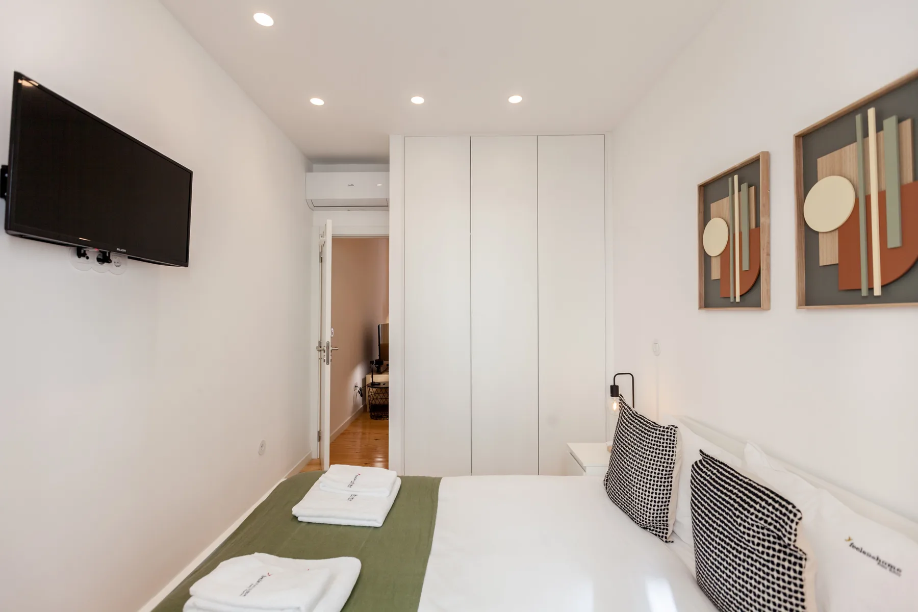 A bedroom with a bed, folded towels, wall-mounted television, and framed artwork.
