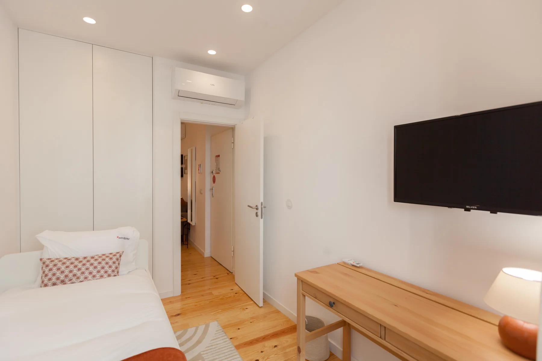 Un dormitorio luminoso con una cama, un televisor de pared y un escritorio de madera.
