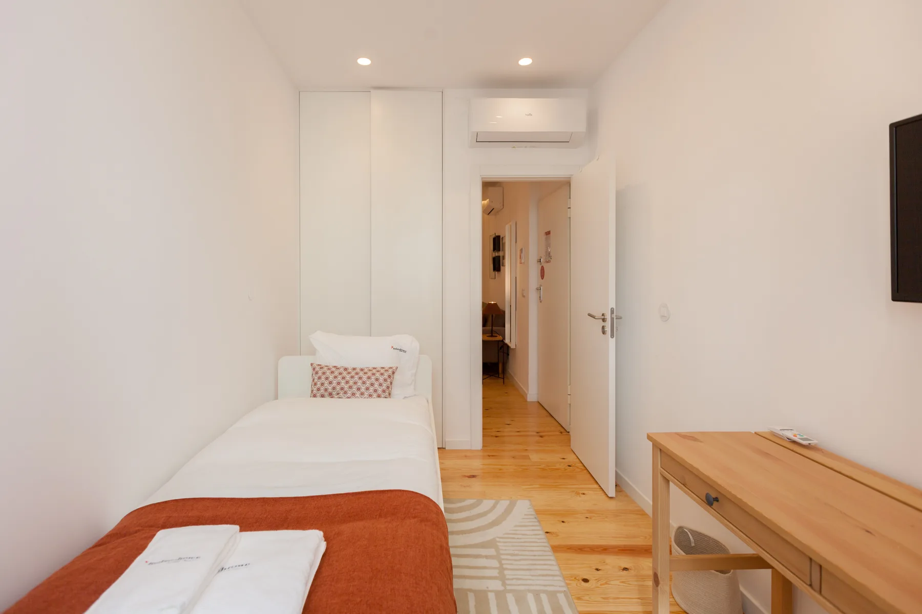Un dormitorio con una cama individual, un escritorio de madera y una puerta hacia el pasillo.