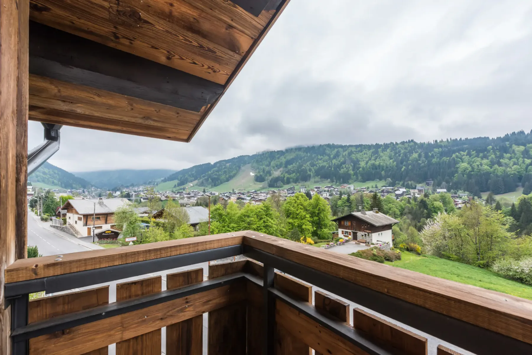 A wooden balcony overlooks a green valley with houses and wooded hills.