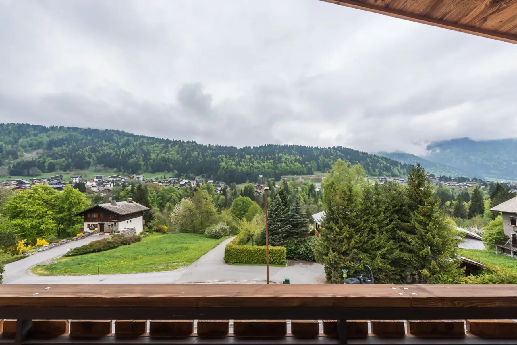 A wooden balcony overlooks a green valley, scattered buildings, and forested hills.