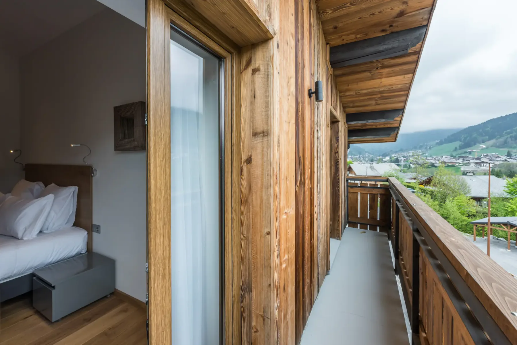 A narrow wooden balcony runs alongside a bedroom, overlooking a green mountain valley.