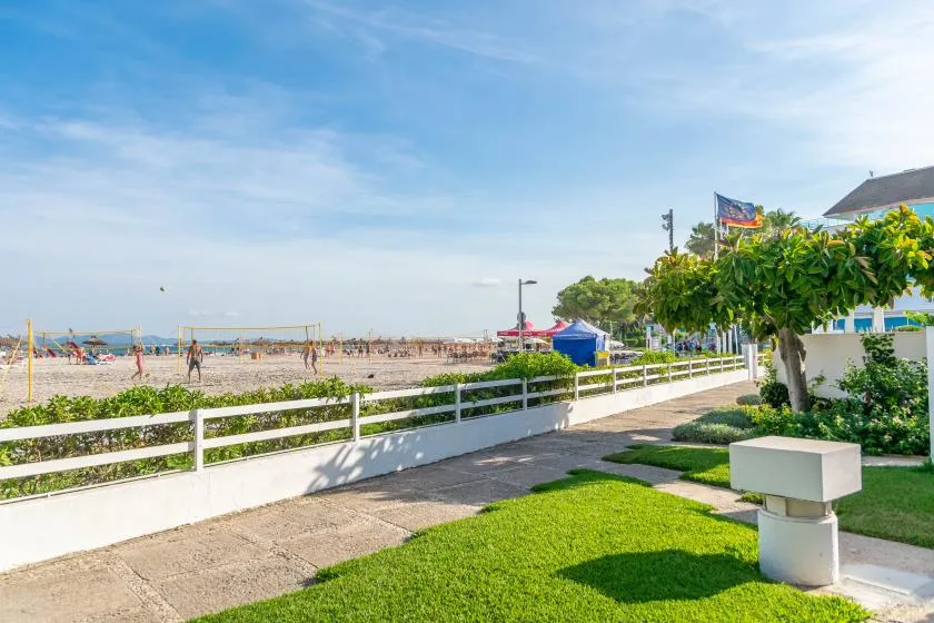 A sunny beachfront walkway runs beside a white fence, with the beach visible beyond.