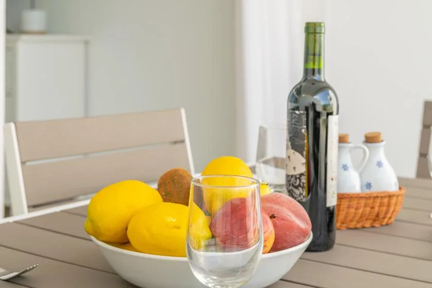A bowl of fruit and a glass sit on a dining table beside a wine bottle.