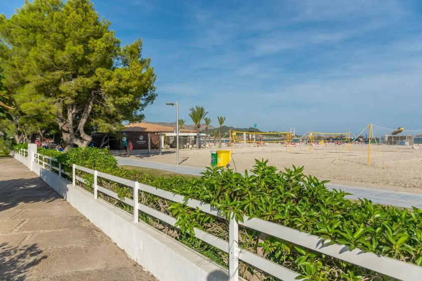 A sandy beach volleyball area sits beside a paved path and leafy greenery.
