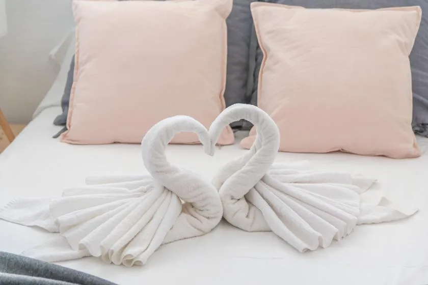 A bed with pale pink pillows and two towels arranged in heart shapes.