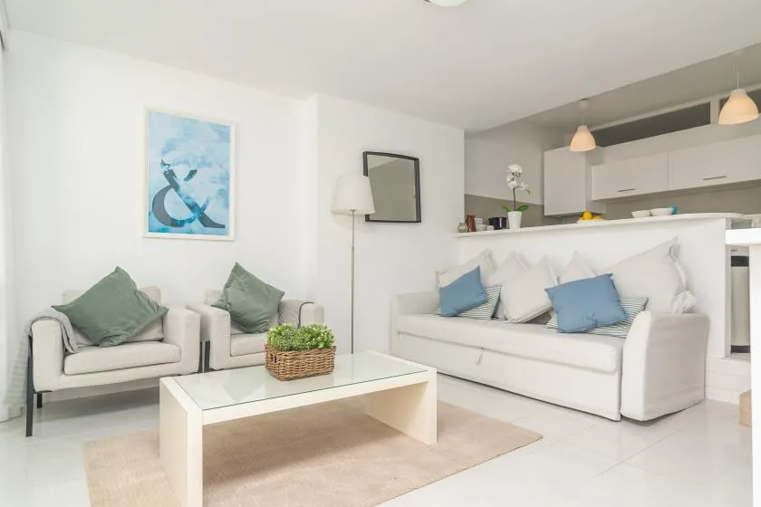 A bright living room with seating around a coffee table and an open kitchen behind it.