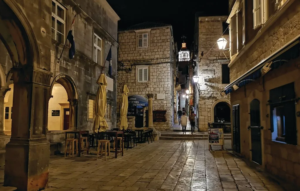 A narrow stone-paved street runs between old buildings with outdoor seating at night.