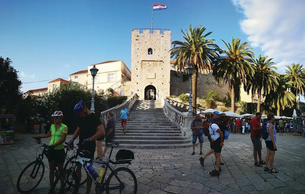 A stone tower stands above a broad outdoor staircase, with palm trees and pedestrians nearby.