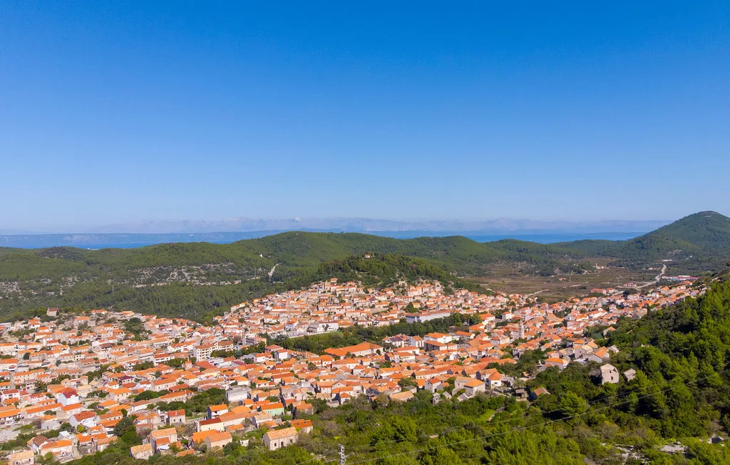 A hillside town with red-tiled roofs spreads across a green valley beneath a clear sky.