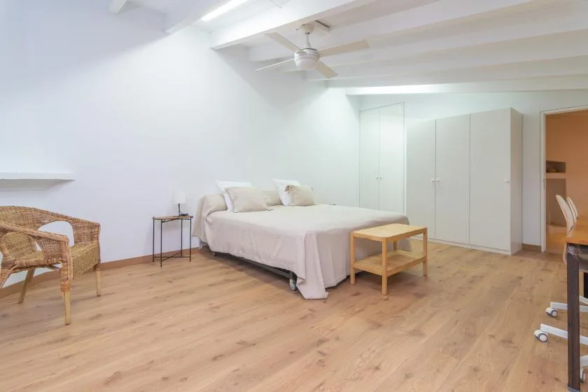 A bright bedroom with a bed, pale walls, and light wood flooring.