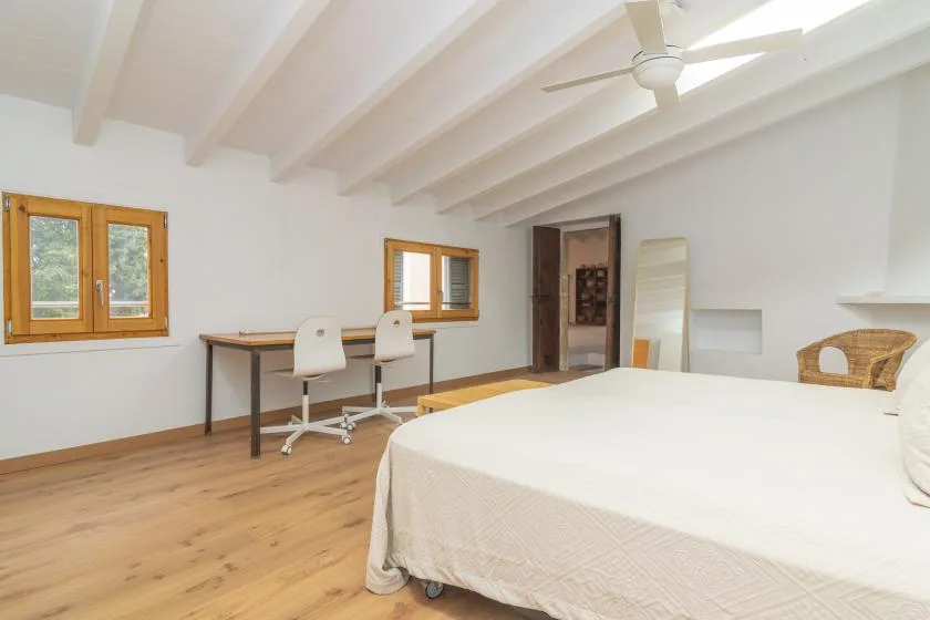A bright bedroom with a bed, desk, and windows beneath a beamed ceiling.