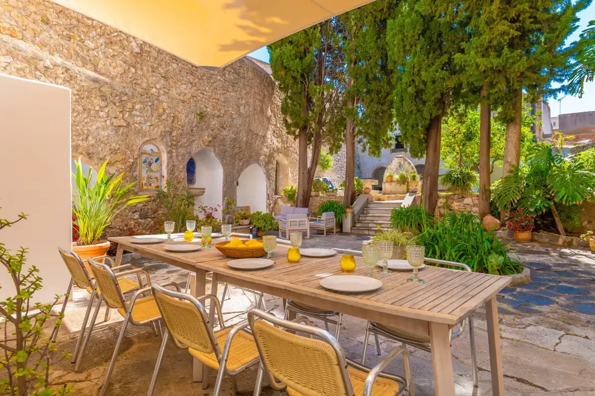 An outdoor dining area with a long table and chairs beneath leafy trees.