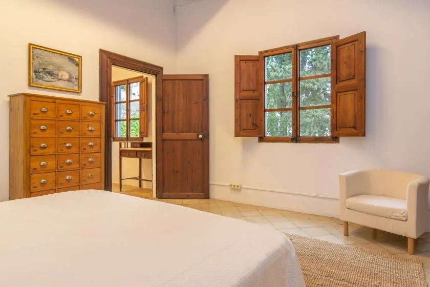 A bedroom with a bed, wooden dresser, and windows with open shutters.