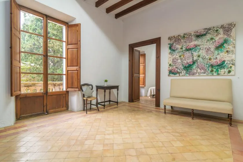 A bright room with tall wooden windows, simple seating, and a colorful wall painting.