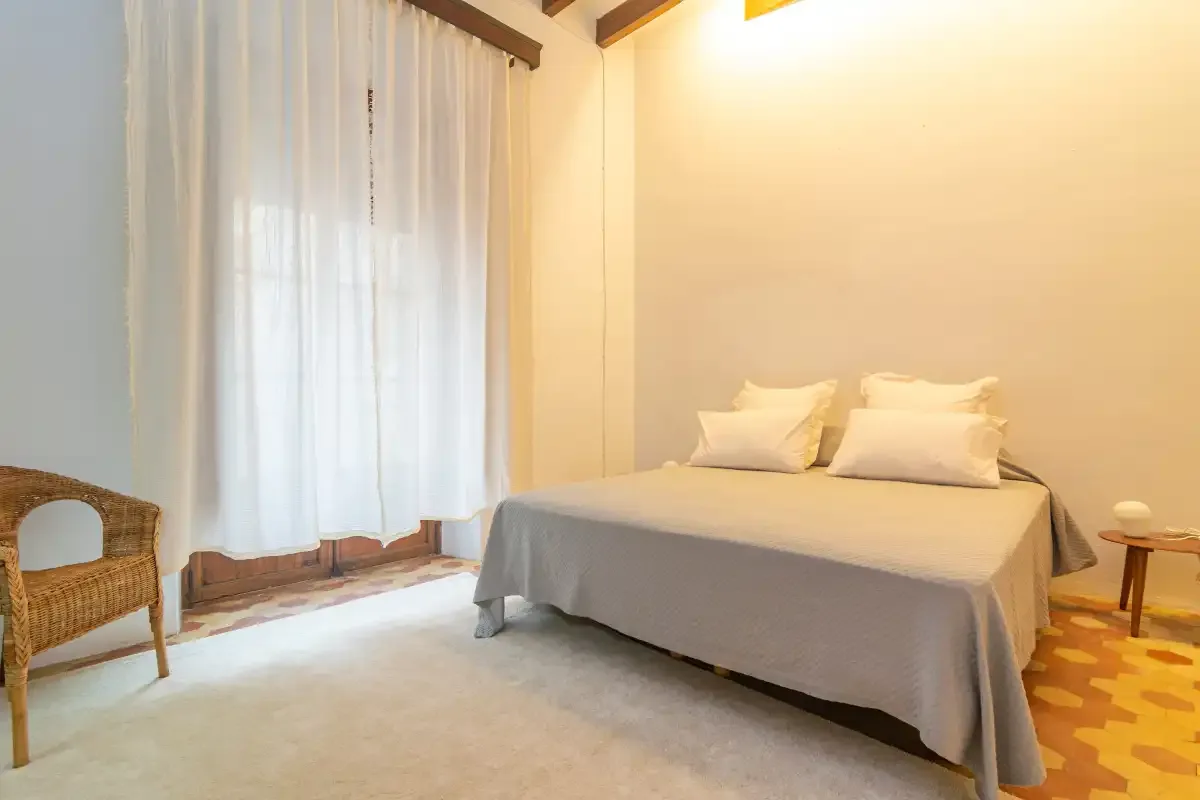 A bedroom with a neatly made bed beside sheer curtains.