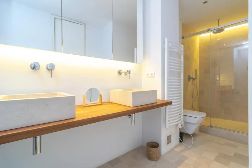 A bright bathroom has a long vanity with two sinks and a glass shower.