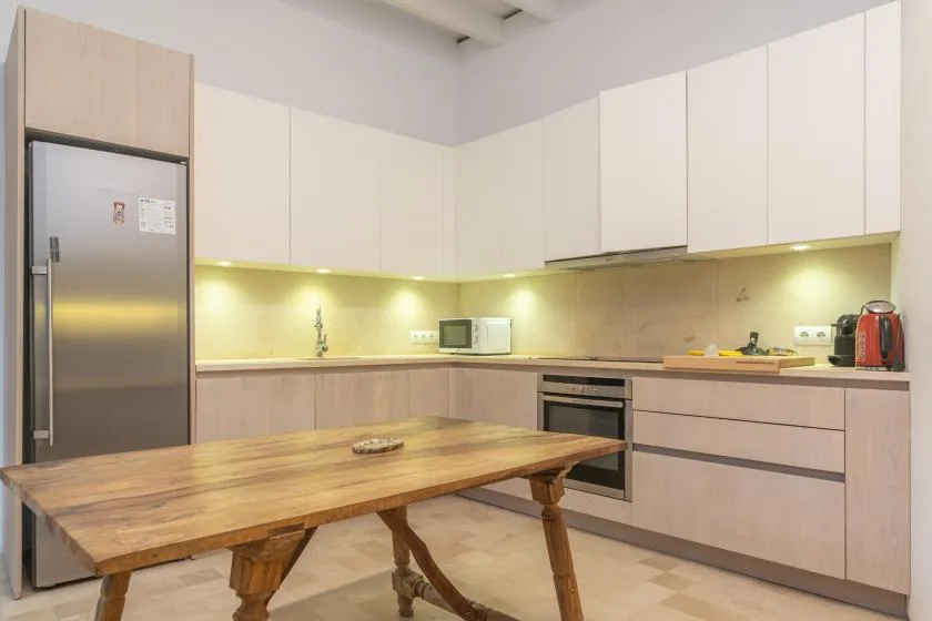 A bright kitchen with pale cabinetry, a stainless-steel refrigerator, and a wooden dining table.