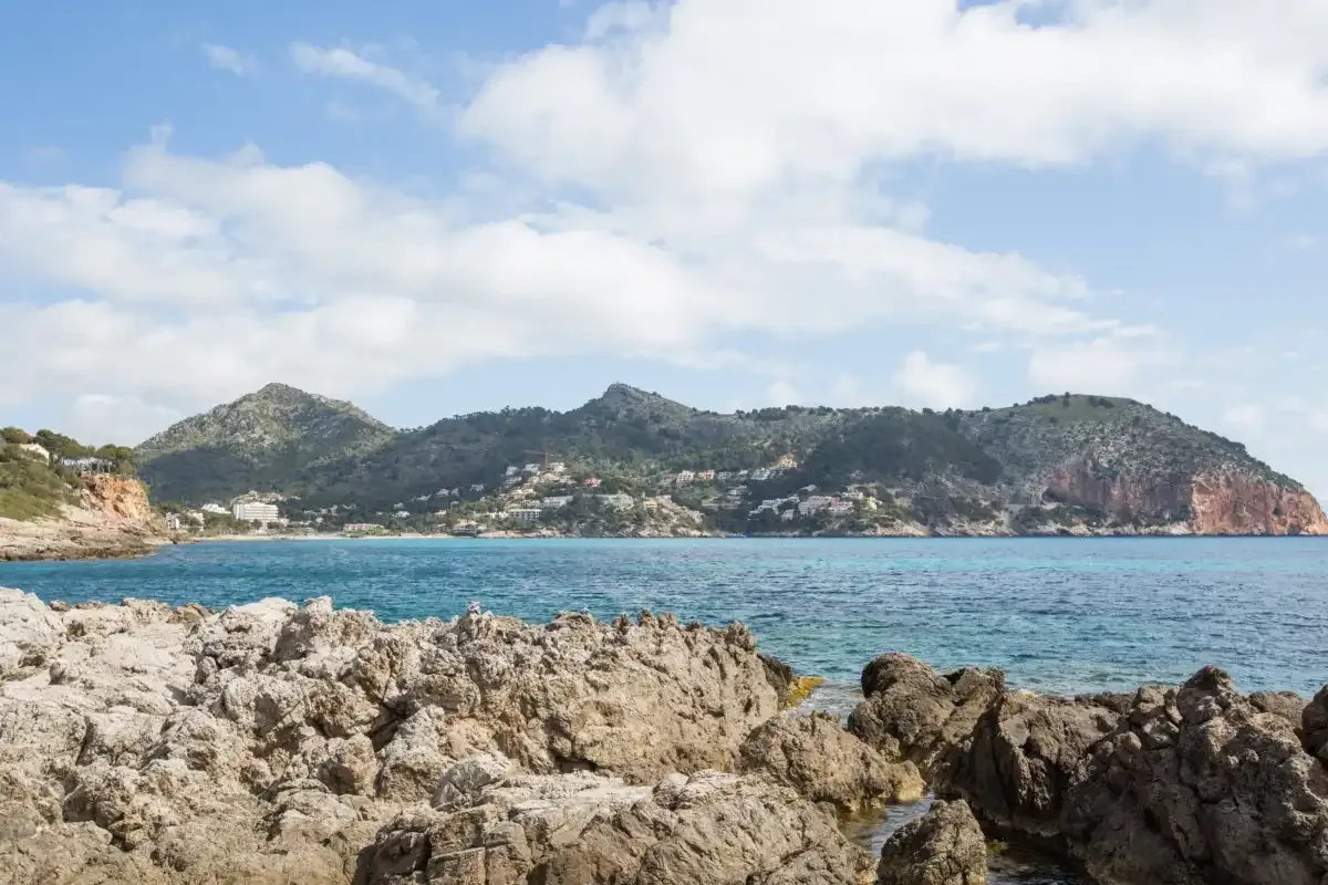 A rocky shoreline overlooks blue water and hills beneath a partly cloudy sky.