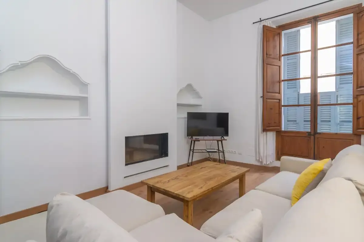 A living room with seating, a coffee table, and a television beside tall windows.