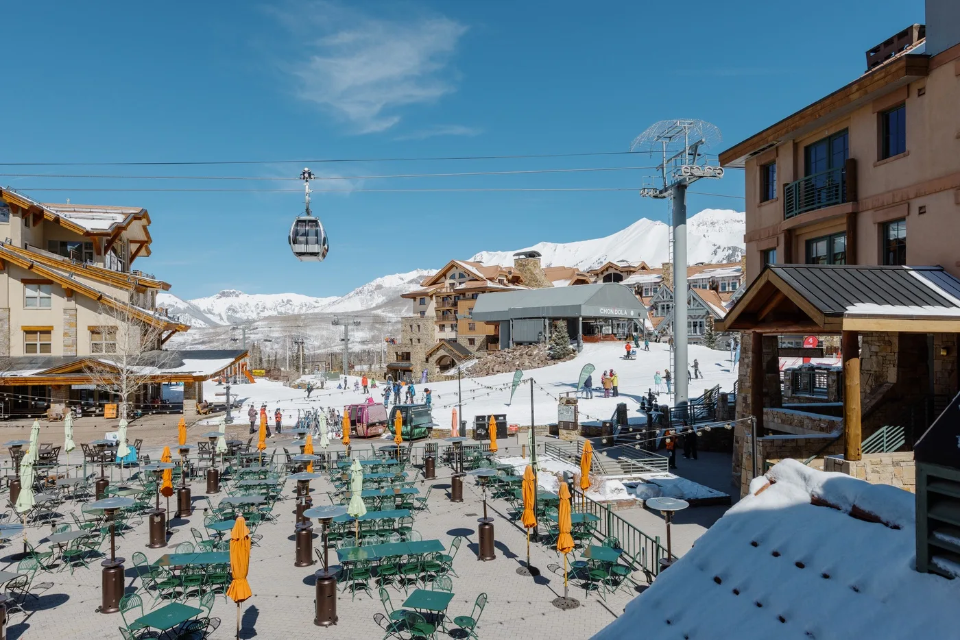 A snow-covered outdoor seating area sits among buildings beneath a mountain gondola.