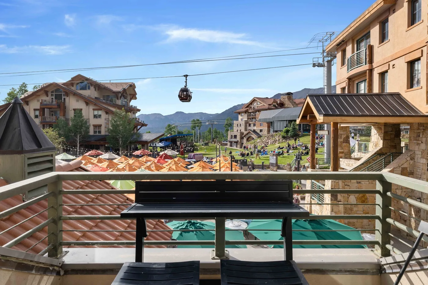 A balcony overlooks resort buildings, outdoor seating, and mountains in the distance.
