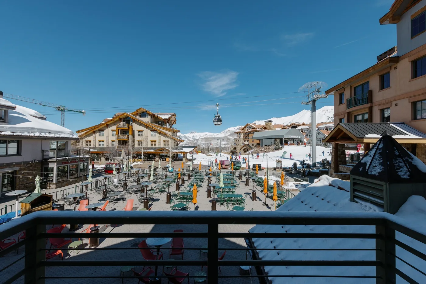 A snowy mountain resort plaza with outdoor seating and a cable car overhead.