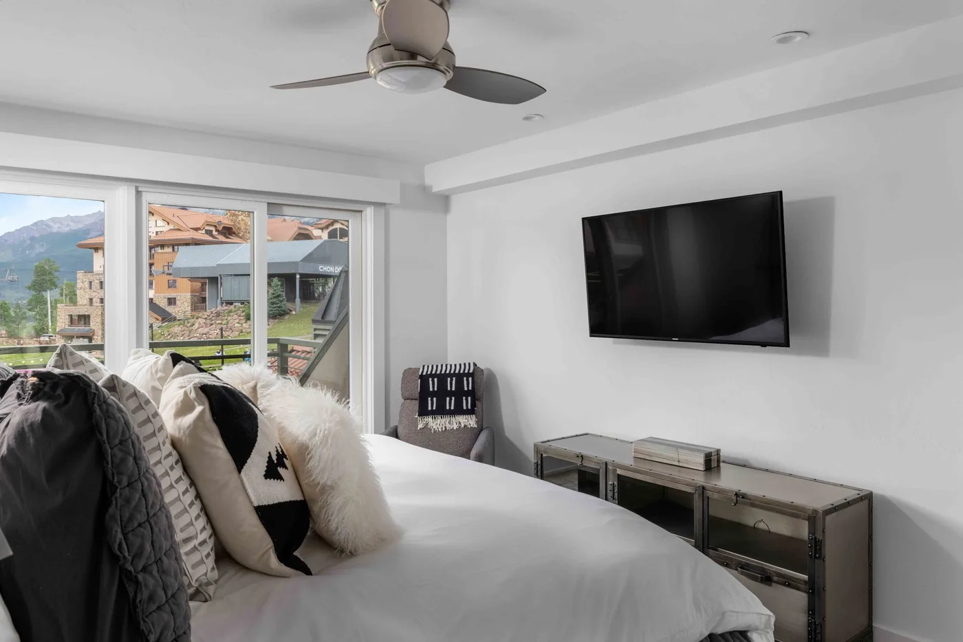 A bright bedroom with a bed beside large windows and a wall-mounted television.