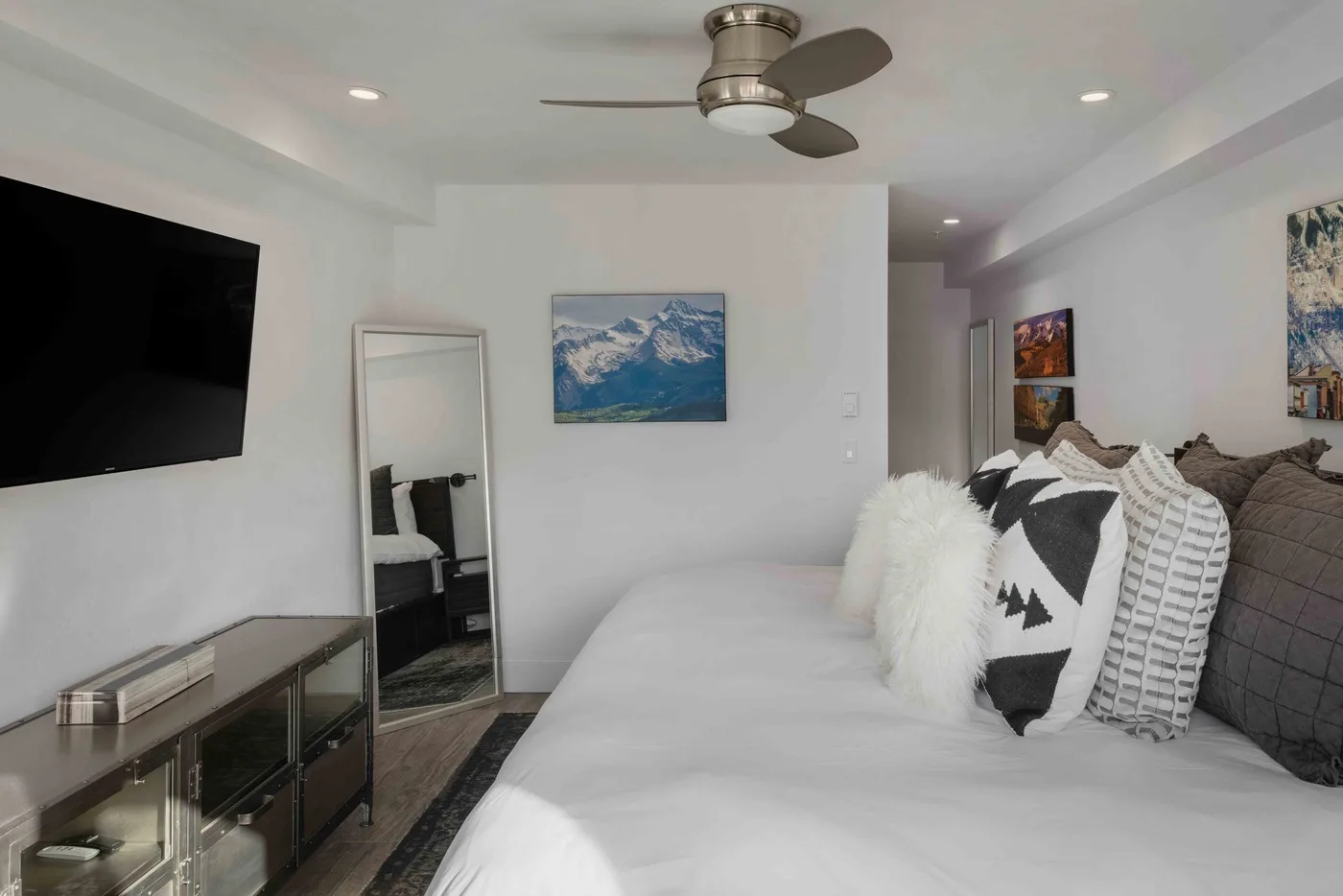 A bright bedroom with a bed, wall-mounted television, and ceiling fan.