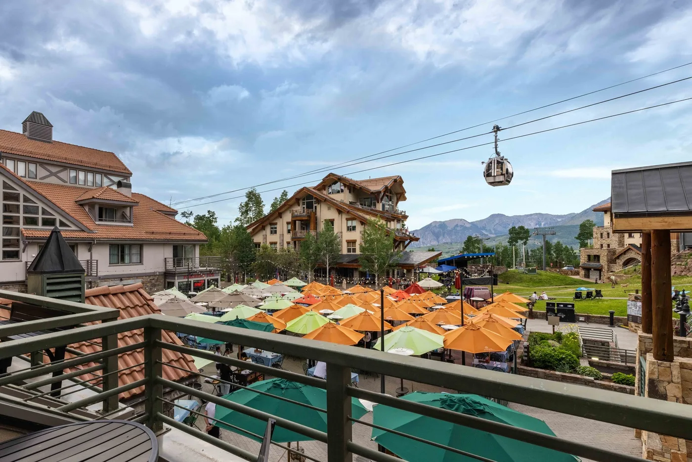 A mountain resort courtyard with colorful umbrellas, surrounding buildings, and a cable car overhead.