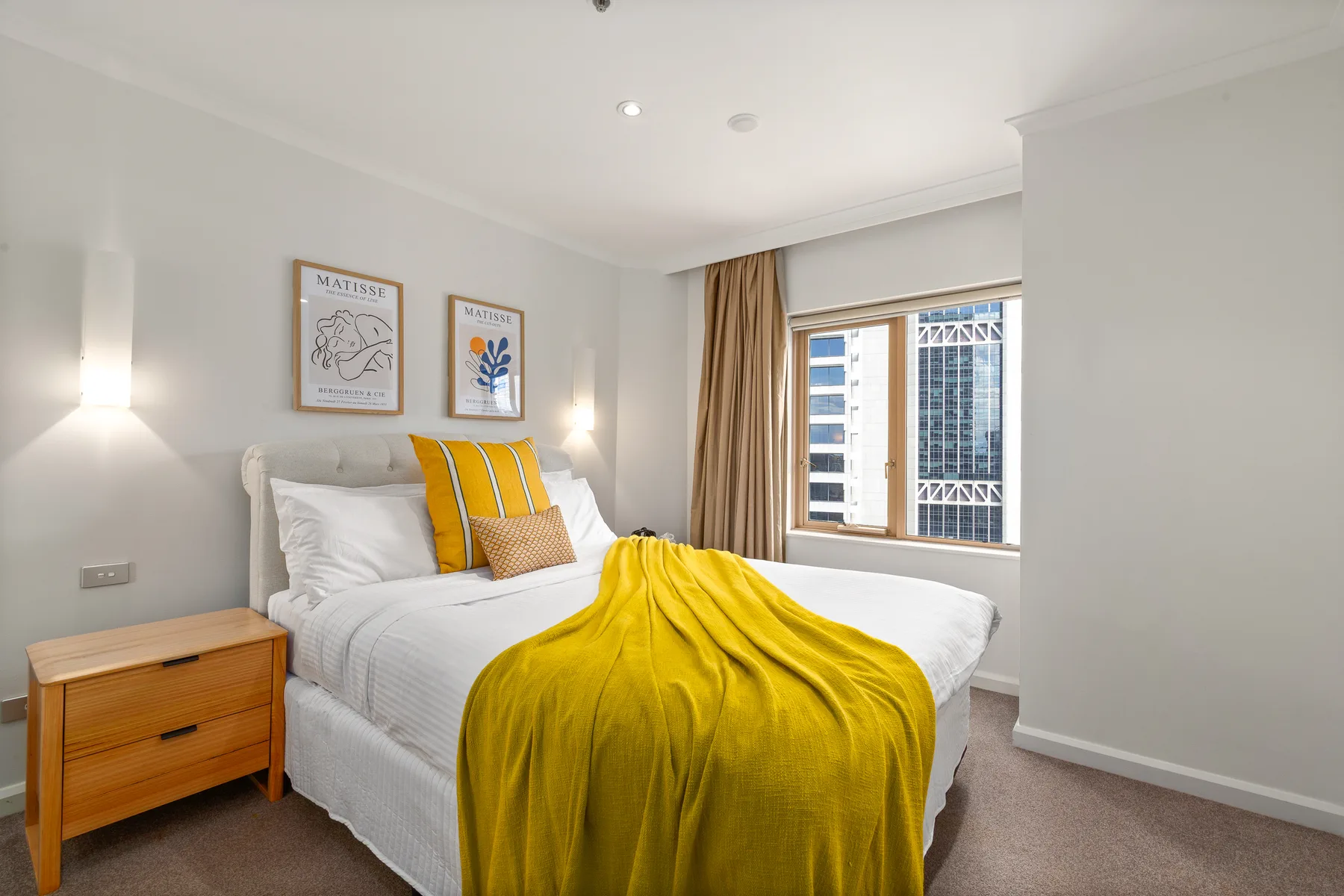 Un dormitorio luminoso muestra una cama con ropa de cama blanca y una manta amarilla junto a una ventana.