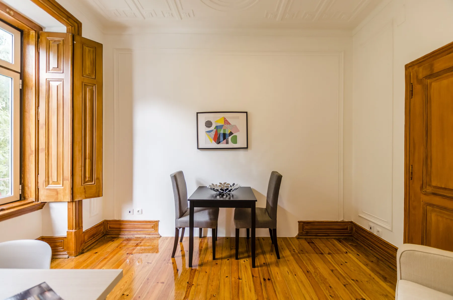 Un comedor con una mesa y sillas está junto a ventanas altas de madera.