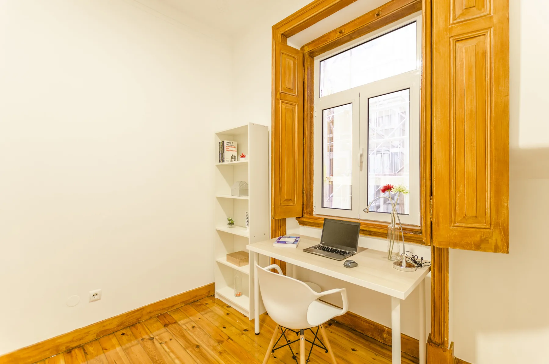 Un escritorio pequeño y una silla están junto a una ventana alta en una habitación luminosa.