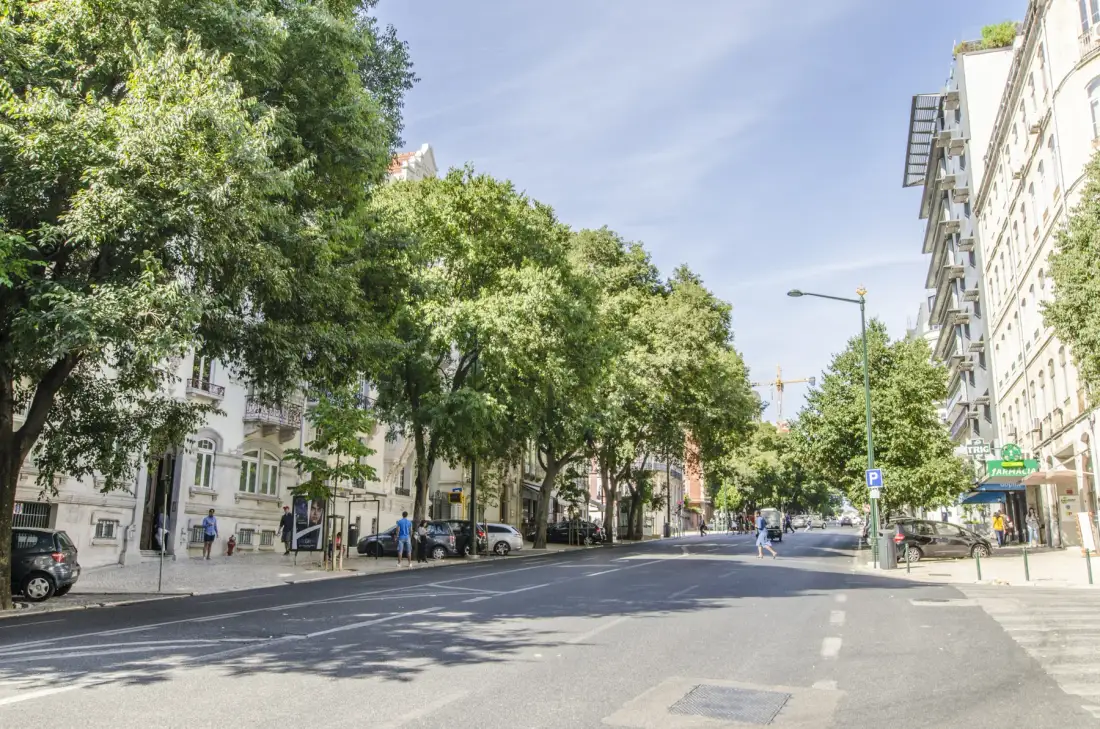 Una calle urbana arbolada con edificios, coches estacionados y peatones.